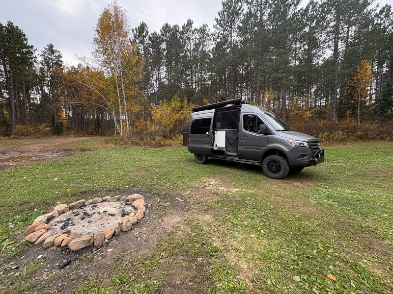  ideal for RV rental adventures. Features off-road exterior and functional interior with beds.|Autumn rv rental adventure: a gray camper van parked by a fire pit in a colorful fall forest setting.|Gray Storyteller Overland rv rental camper van on grass. Side door open — photo 2