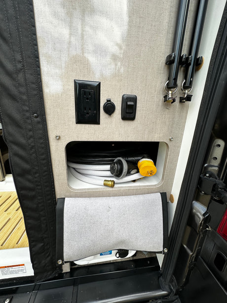 RV rental: Organized utility panel with AC outlets, water hookups, electrical adapters, and stored awning poles.