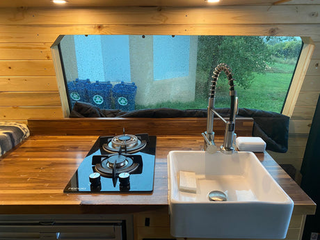 Cozy rv rental campervan kitchen with wood counters, stove, sink, and rainy window view of recycling bins and green landscape.