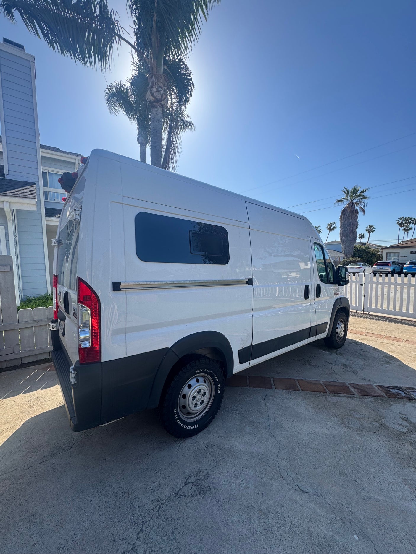 White Ram ProMaster camper van, ideal for rv rental, parked on a sunny concrete driveway with palm trees.