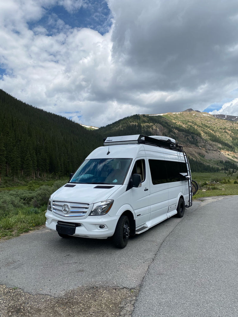  parked amidst tall pines and green grass with a rocky mountain backdrop.|White Mercedes-Benz Sprinter RV rental on a mountain road — photo 3