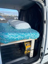 RV rental interior: a bed with tropical sheets and pillows, black curtains, and a yellow CAT portable power station inside a camper van.