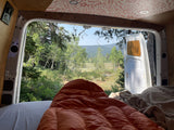 Inside an RV rental, a comfy sleeping bag faces open van doors revealing a lush forest and distant mountains.
