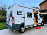 White camper van **rv rental** with open side door, interior lights, & awning extended. Rear ladder visible.