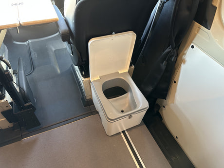 An RV rental's practical interior: A grey portable composting toilet, lid open, is placed next to the swivel passenger seat.