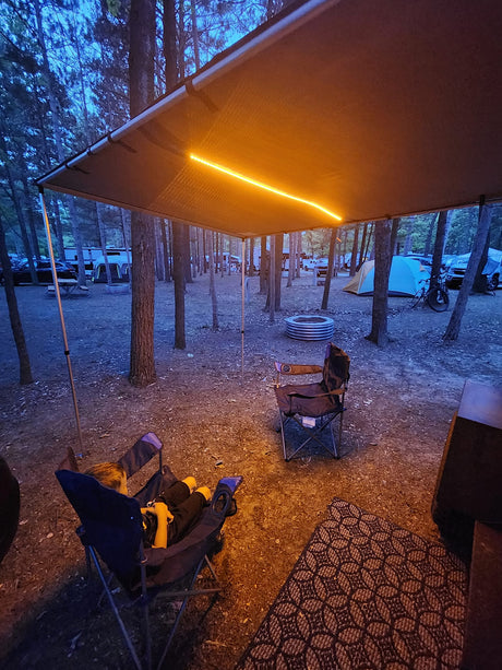 Cozy evening at an RV rental campsite. Warm LED lights illuminate the awning over chairs, with other campers in the background.