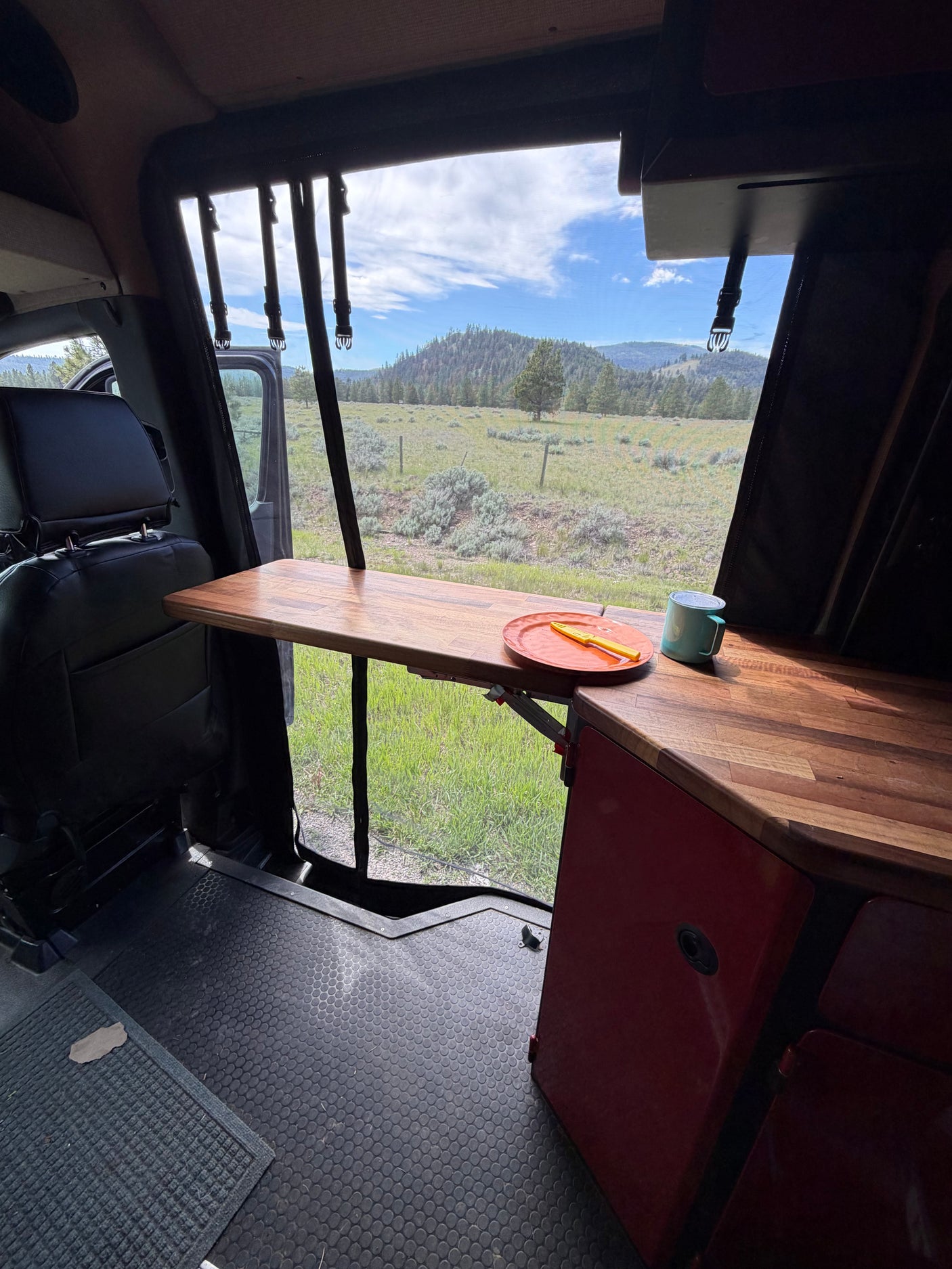 Inside an RV rental, a wooden table with a plate and mug faces a scenic mountain landscape through an open door.