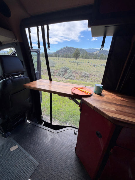 Inside an RV rental, a wooden table with a plate and mug faces a scenic mountain landscape through an open door.