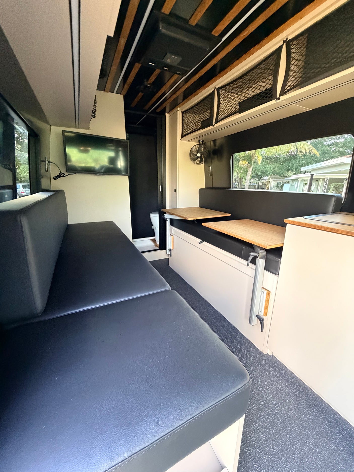 Spacious RV rental interior with black cushioned seating, adjustable tables, TV, and bathroom access. Ideal for comfortable travel.