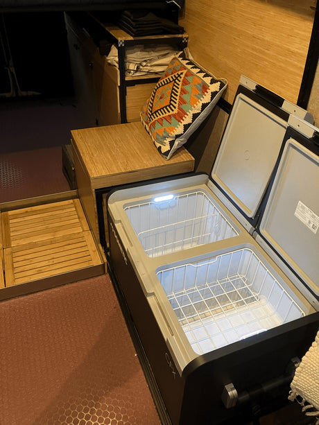 Inside an RV rental: a Dometic portable fridge/freezer, open to reveal wire baskets, next to wood storage & pull-out tray.