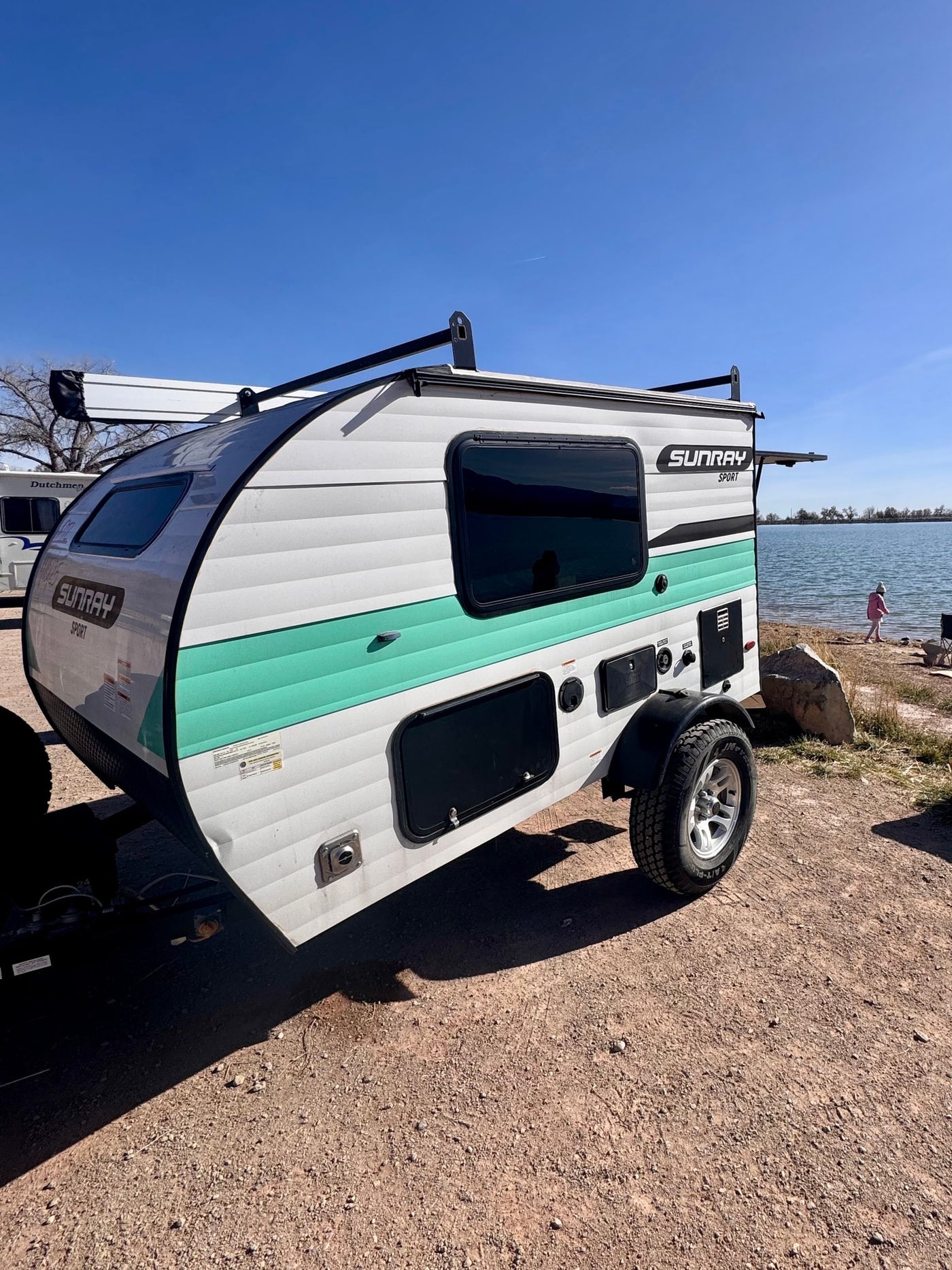 This white & teal Sunray Sport teardrop rv rental is parked lakeside. A child plays nearby on a sunny day. — photo 10