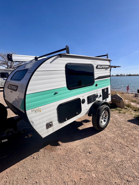 This white & teal Sunray Sport teardrop rv rental is parked lakeside. A child plays nearby on a sunny day.