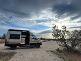 Adventure-ready rv rental camper van parked on a gravel lot with its door open, set against a dramatic cloudy sky and desert landscape.