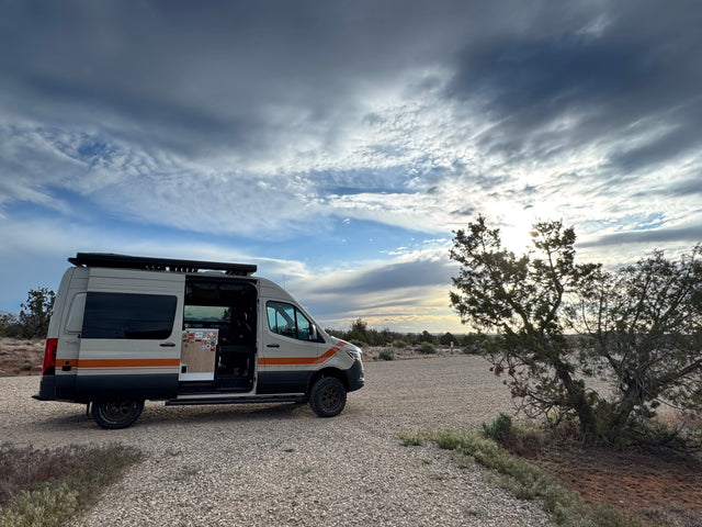 Adventure-ready rv rental camper van parked on a gravel lot with its door open, set against a dramatic cloudy sky and desert landscape.
