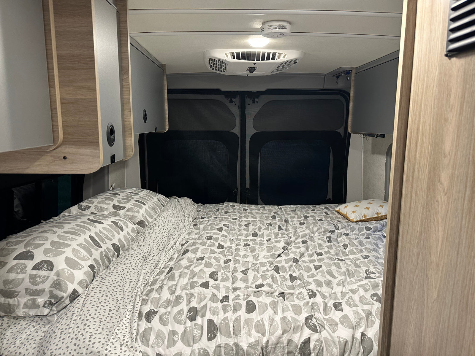 RV rental's cozy bedroom features patterned bedding, sleek overhead cabinets, and dark rear window screens for privacy.