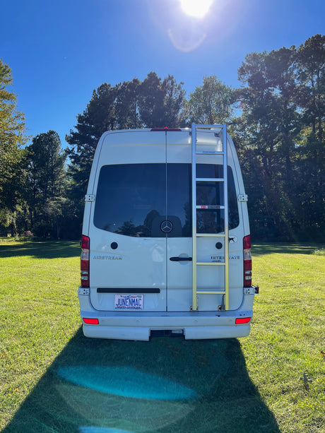 A white Airstream Interstate RV rental, seen from the rear with a ladder, sits on a sunny green lawn surrounded by trees.