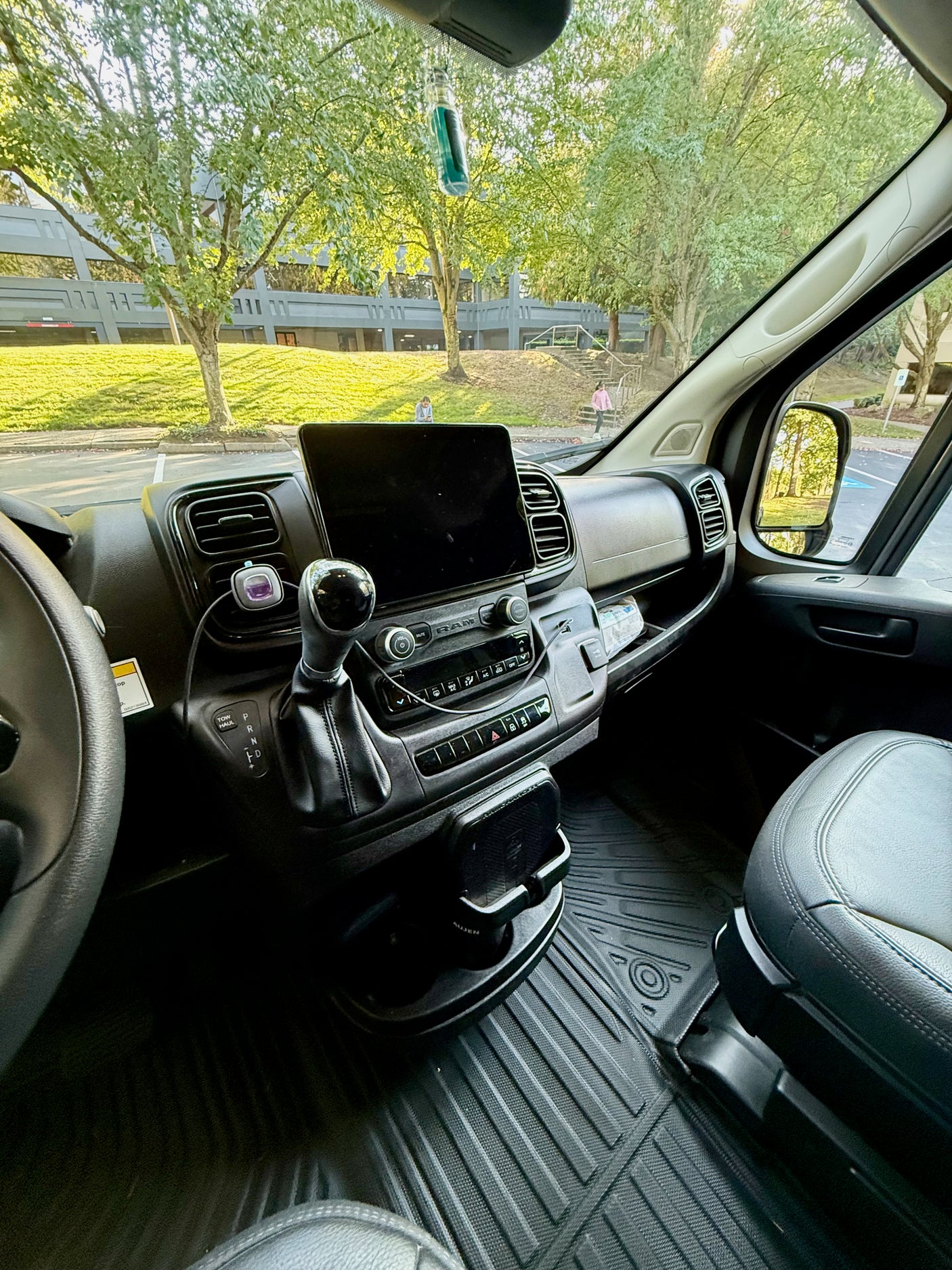 Interior view of an RV rental's cockpit, featuring a tablet on the dash, steering wheel, and a view of trees and a building.