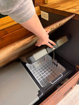 Hand opening a compact refrigerator/freezer with a basket, built into custom wooden cabinetry for an RV rental.