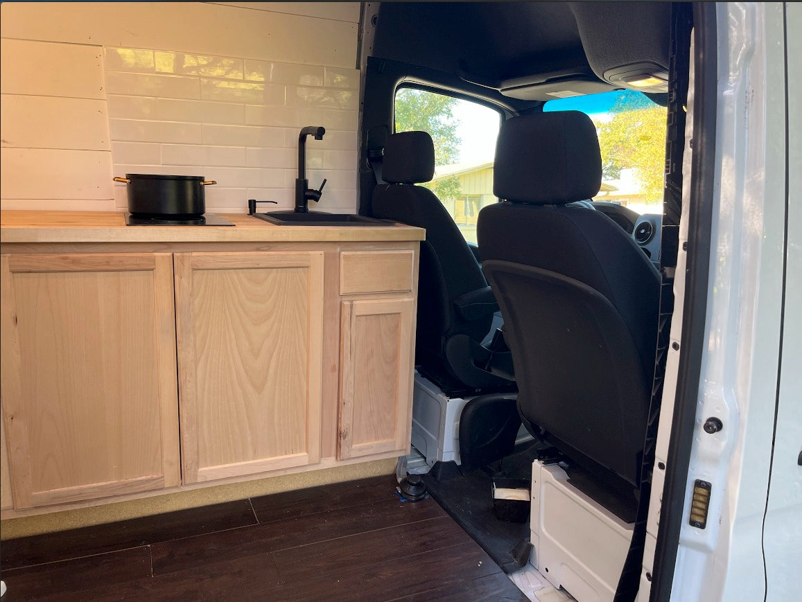 Inside an RV rental: a modern kitchen with wood cabinets, black sink, and cooktop. View of front seats.