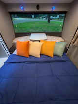Cozy RV rental interior with a blue comforter, colorful pillows, and a large window view of a campsite.