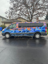 This vibrant rv rental van, adorned with abstract blue, orange & purple art, is parked on a wet residential street, ready for travel.