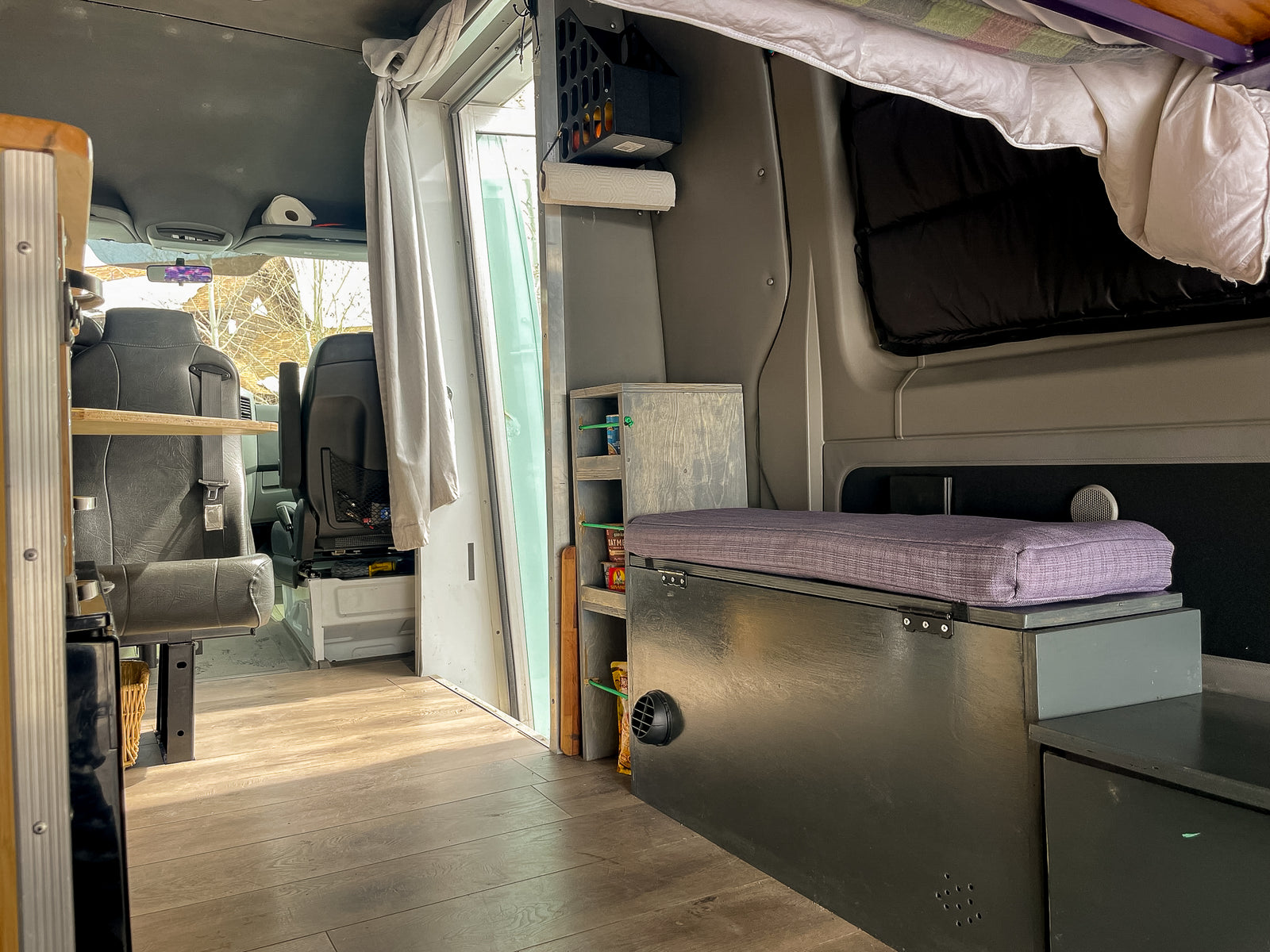 Inside this custom RV rental: wood floor, front seats, kitchen shelf, and a purple-cushioned bench. Ideal mobile living.