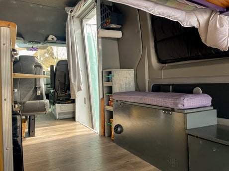 Inside this custom RV rental: wood floor, front seats, kitchen shelf, and a purple-cushioned bench. Ideal mobile living.