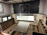 An rv rental camper interior showcasing a cozy dinette with bear and canoe patterned benches, a table, and large window with curtains.