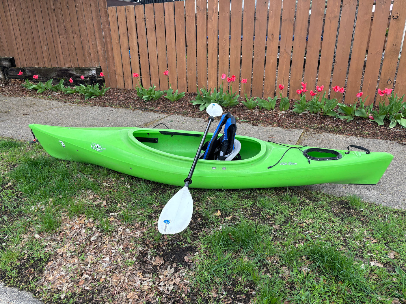 Bright green kayak and paddle on grass by a fence with pink tulips. A serene outdoor scene, perhaps a stop on an RV rental journey.