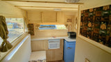 A light-wood rv rental interior with a kitchenette, blue retro fridge, window AC, and a window overlooking greenery.