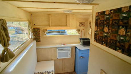 A light-wood rv rental interior with a kitchenette, blue retro fridge, window AC, and a window overlooking greenery.