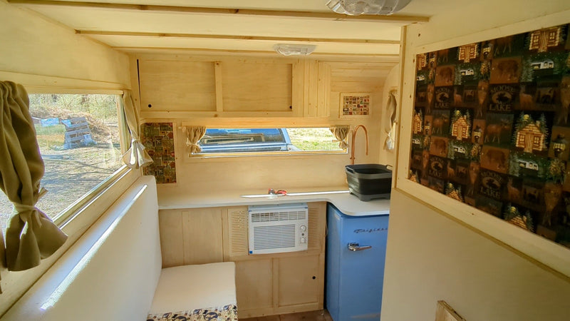 A light-wood rv rental interior with a kitchenette, blue retro fridge, window AC, and a window overlooking greenery. — photo 3
