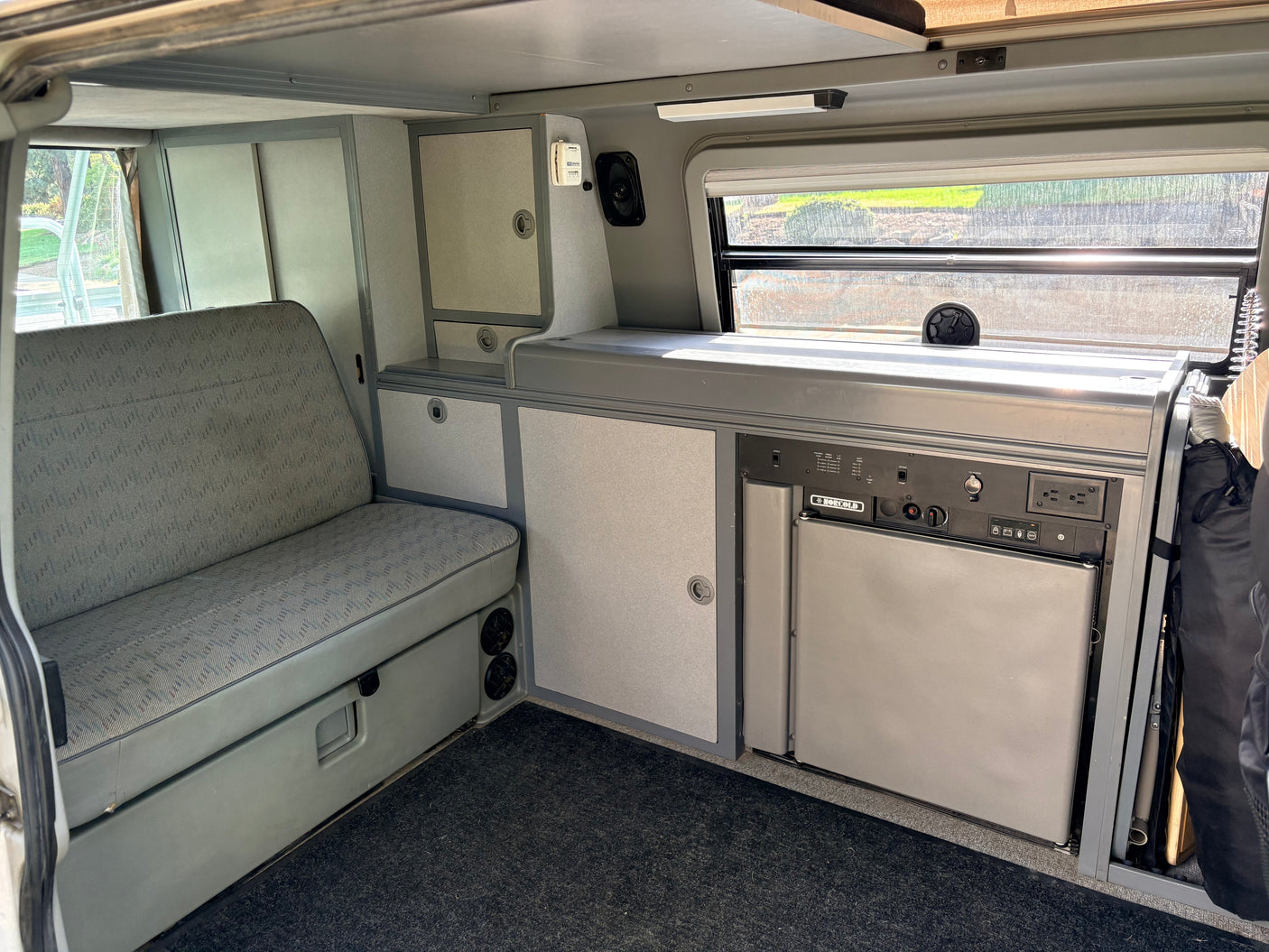 RV rental interior: Compact camper van with a gray upholstered bench, kitchenette, fridge, and control panel.