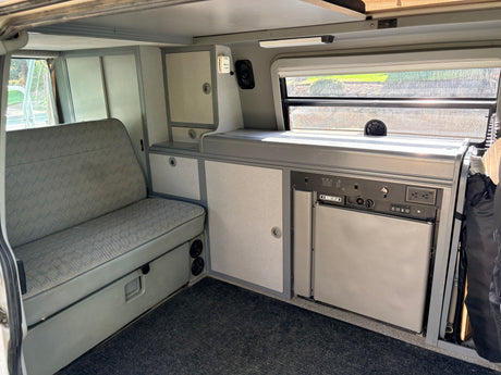 RV rental interior: Compact camper van with a gray upholstered bench, kitchenette, fridge, and control panel.