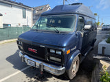 A well-loved blue GMC Vandura high-top van, potentially an rv rental, showing paint wear, parked on a sunny street.