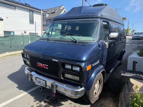 A well-loved blue GMC Vandura high-top van, potentially an rv rental, showing paint wear, parked on a sunny street.