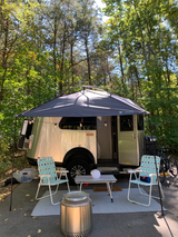 An rv rental camping setup: silver travel trailer, shaded canopy, chairs, table, and fire pit under tall trees.