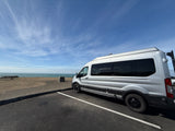 This silver RV rental is parked coastside. A camper van overlooks the vast blue ocean under a vibrant sky, ready for adventure.