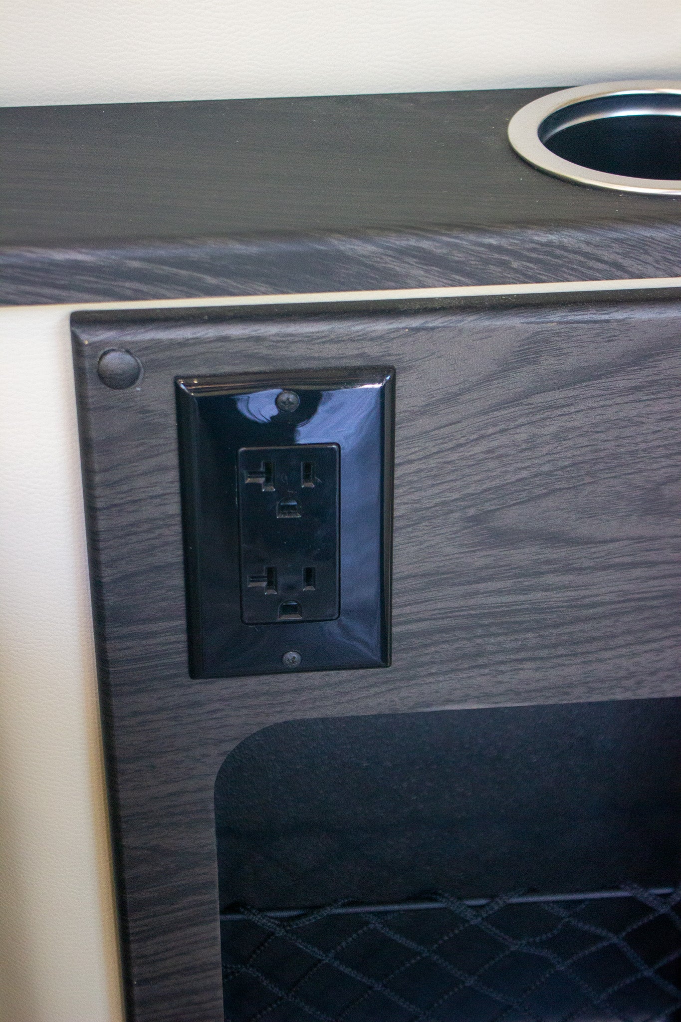 RV rental power and storage: A black electrical outlet, cupholder, and mesh pocket in a dark wood interior.