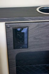 RV rental power and storage: A black electrical outlet, cupholder, and mesh pocket in a dark wood interior.