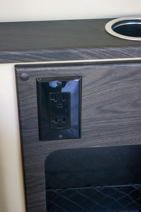 RV rental power and storage: A black electrical outlet, cupholder, and mesh pocket in a dark wood interior.