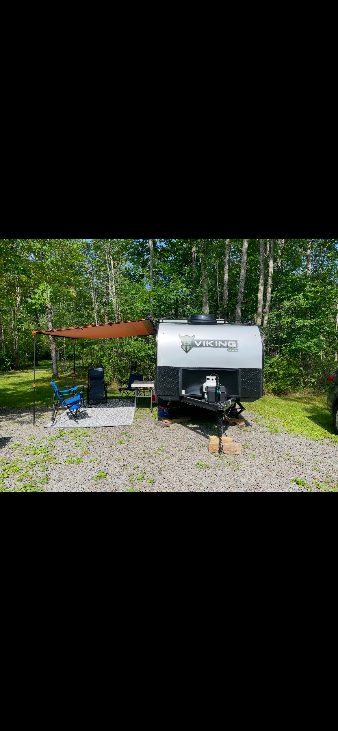 RV rental: Viking Max travel trailer with awning, chairs, and table at a sunny, wooded campsite.