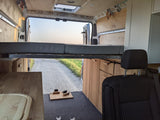 Custom van interior, ideal for an **rv rental**. Features a raised bed, kitchen, and open rear doors revealing a gravel road.