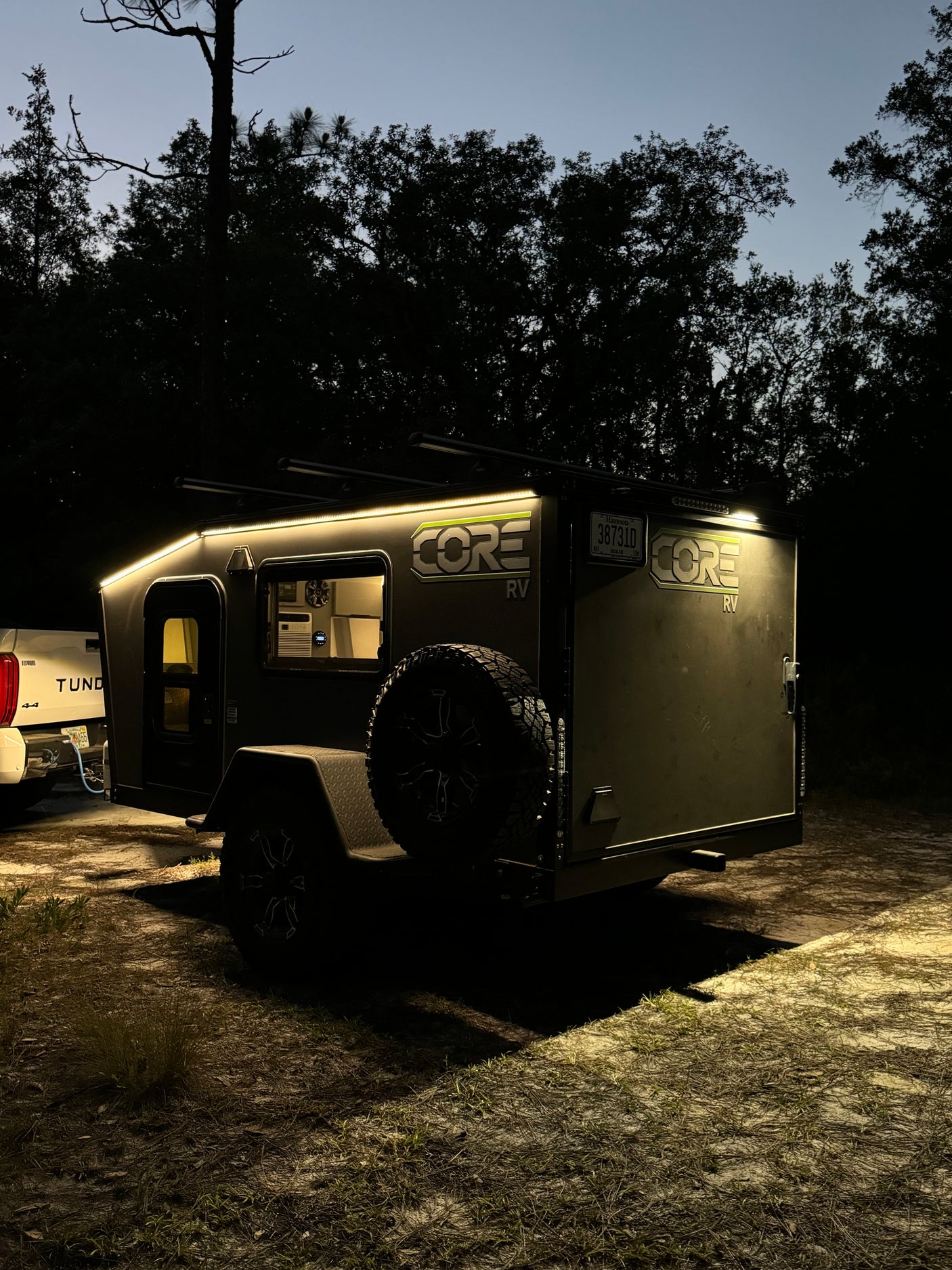 Experience camping with an RV rental! This CORE RV trailer glows at night, parked in a forest campsite with a truck.