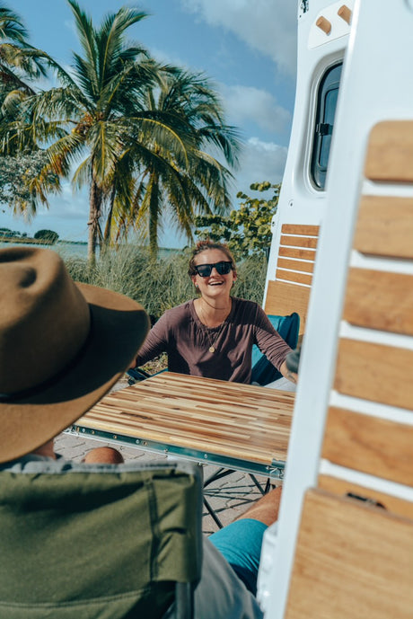 Happy woman smiles at outdoor table next to an RV rental. Palm trees & water in sunny tropical scene.