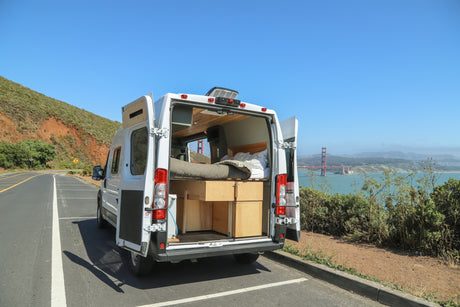 White RV rental camper van with open doors, revealing bed & storage, parked overlooking Golden Gate Bridge.
