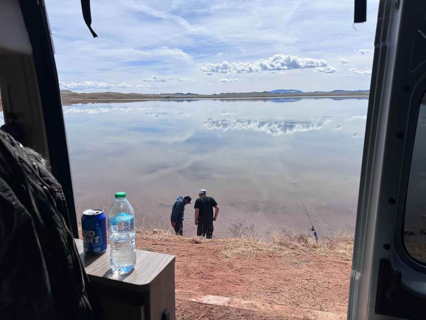 Perfect day for lake fishing from our RV rental. Calm water reflects the sky, two men fish by the shore. Drinks ready inside.