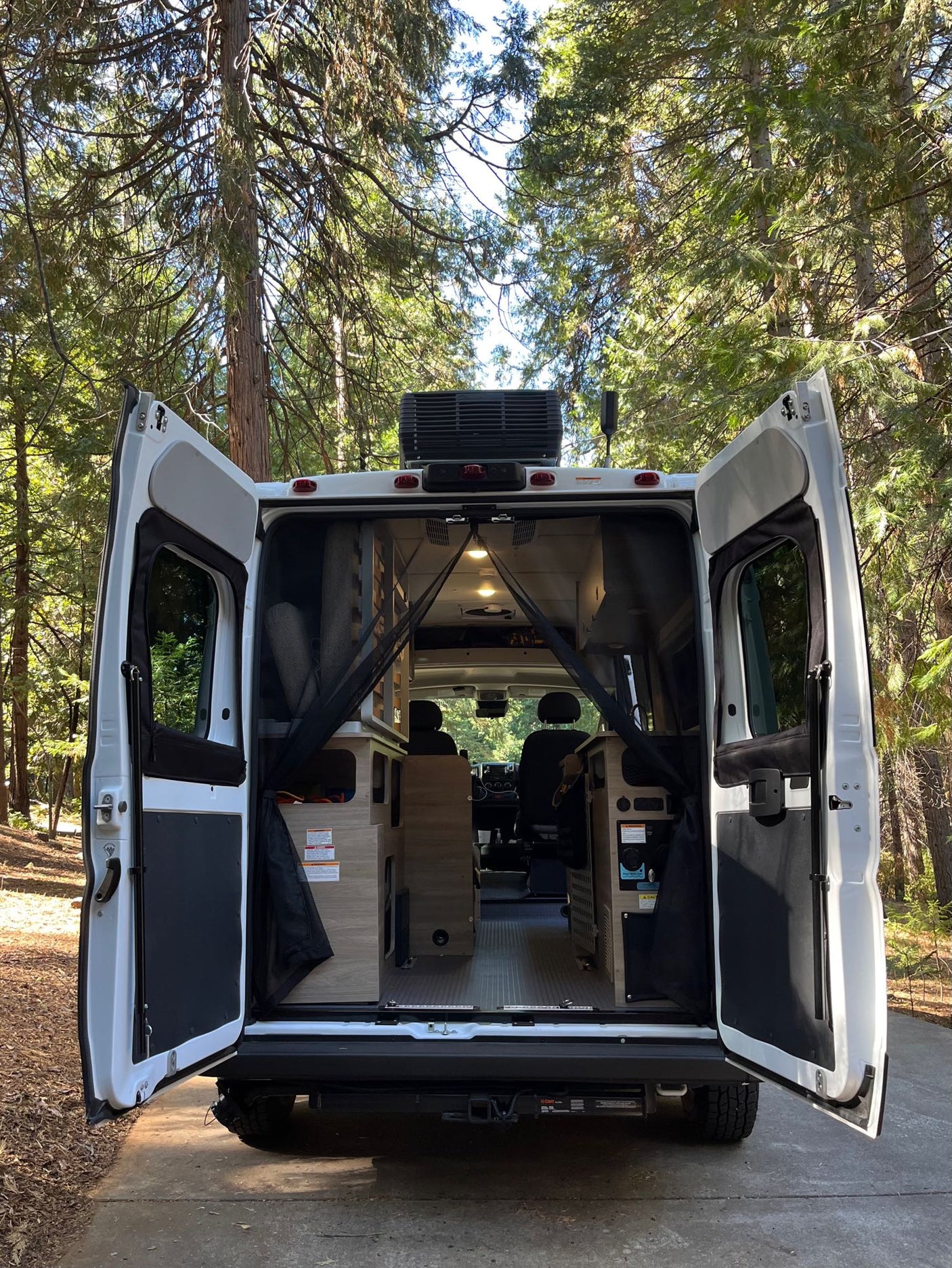 RV rental's rear doors open in a sunny forest, showcasing its cozy interior ready for adventure and travel.