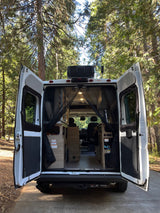 RV rental's rear doors open in a sunny forest, showcasing its cozy interior ready for adventure and travel.