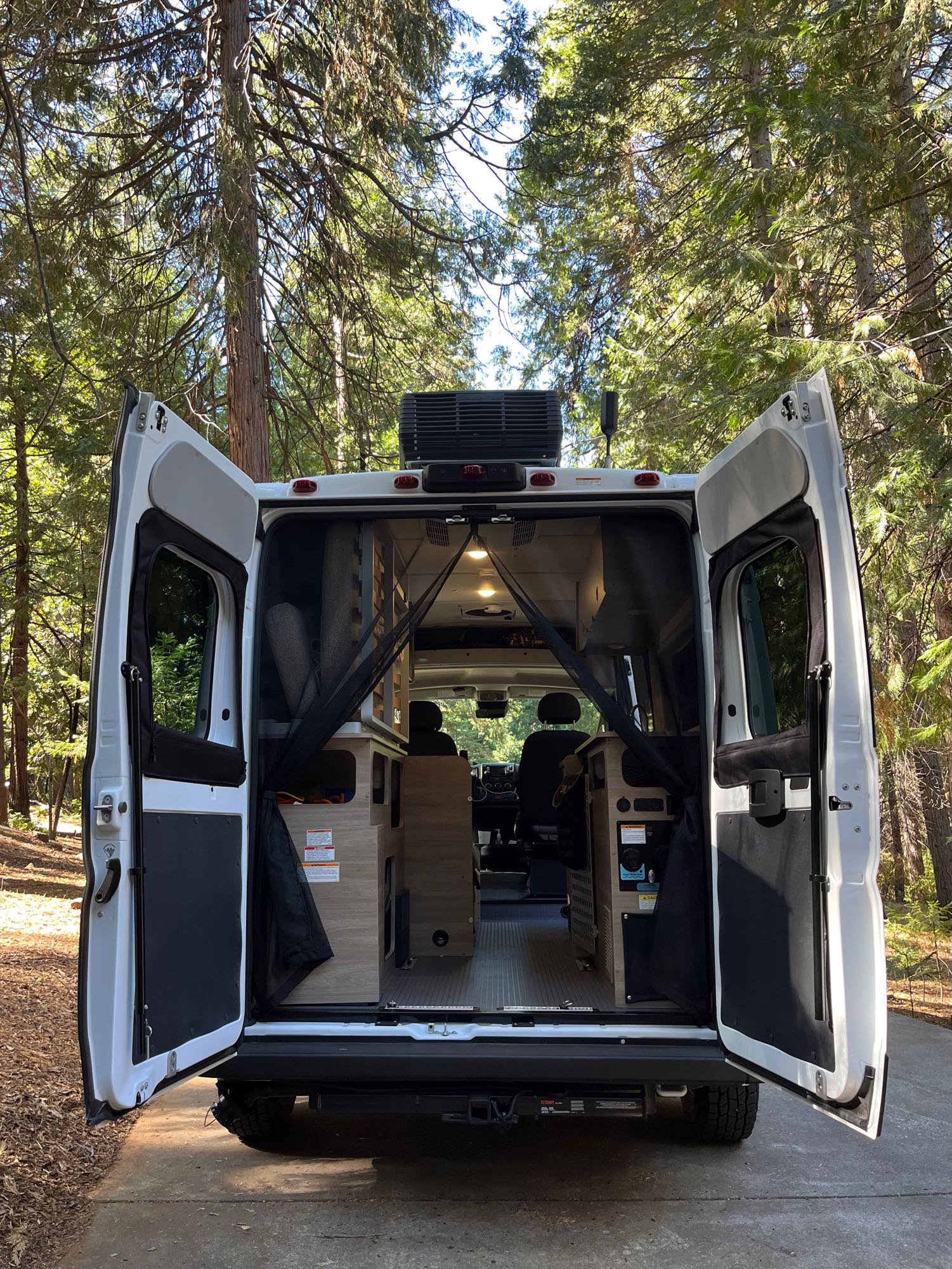 RV rental's rear doors open in a sunny forest, showcasing its cozy interior ready for adventure and travel.